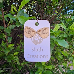 May include: A pair of wooden sloth earrings with a natural wood finish. The earrings are shaped like a sloth's face and have a small hole for the earring post. The earrings are displayed on a white card with the text "Sloth Creations".