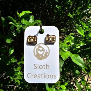 May include: A pair of wooden camera earrings with a light brown wood base and a yellow camera design. The earrings are displayed on a white card with a sloth logo and the text "Sloth Creations".