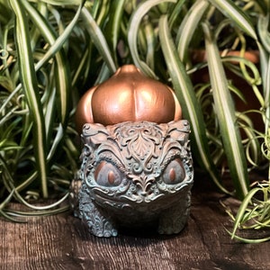 May include: A detailed, ornate, green and copper-colored figurine of a Pokemon character, Squirtle, with a copper-colored shell. The figurine is partially obscured by a potted plant with long, green and white striped leaves.