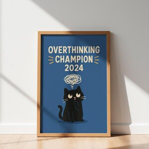 May include: Framed art print with a black cat illustration and the text "OVERTHINKING CHAMPION 2024" on a blue background. The cat has a thought bubble above its head. The print is in a wooden frame and displayed on a wooden floor.