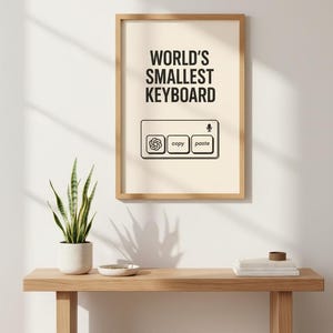 Puede incluir: Una impresión enmarcada con el texto "WORLD'S SMALLEST KEYBOARD" encima de un gráfico de un teclado con los botones "copy" y "paste". La impresión se muestra en un banco de madera clara con una planta en maceta y objetos decorativos.