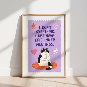 May include: A framed art print with a black and white cat illustration. The print reads "I don't overthink. I just have epic inner meetings." The background is light purple with a heart and star. The frame is wooden.