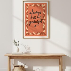 May include: A framed art print with the text "always kiss me goodnight" in a handwritten style. The print has a burnt orange background with a decorative leaf border. The frame is a light wood color.