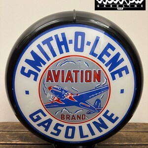 May include: A round, black-rimmed Smith-O-Lene Aviation Gasoline sign with a white face, blue lettering, and a central graphic of a blue and red airplane. The text reads Smith-O-Lene Aviation Brand Gasoline, with a Hot Rod Gasoline LLC logo above.