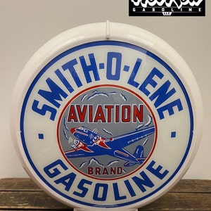 May include: A round white Smith-O-Lene Aviation Gasoline sign with blue and red text and a graphic of a blue airplane. The sign features the text Smith-O-Lene, Aviation, and Gasoline, with a white border and a white base stand for display.