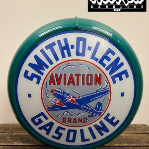 May include: A round, teal-rimmed Smith-O-Lene Aviation Gasoline sign featuring a central graphic of a blue and red airplane against a grey background. The text reads Smith-O-Lene Aviation Brand Gasoline in blue and red lettering on a white face.