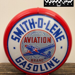 May include: A round, red-rimmed Smith-O-Lene Aviation Brand Gasoline sign with a white center. The graphic features a blue and red airplane flying through clouds. The text Smith-O-Lene is in blue, Aviation is in red, and Gasoline is in blue.