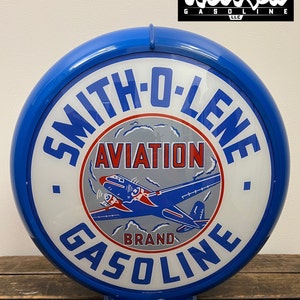 May include: A vintage-style Smith-O-Lene Aviation Gasoline sign. The sign is round with a blue border and features the text "SMITH-O-LENE AVIATION GASOLINE BRAND" in white and red. A blue airplane graphic is in the center.