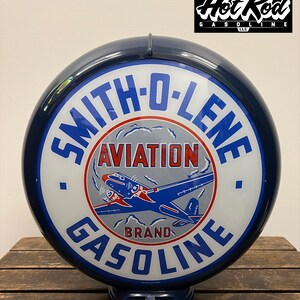 May include: A round, dark blue gas pump globe featuring the text SMITH-O-LENE AVIATION BRAND GASOLINE. The center graphic displays a blue and red airplane flying through white clouds against a light gray background, all encased in a glossy frame.