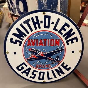 May include: A circular metal sign featuring a white background with a dark blue border. The text reads SMITH-O-LENE AVIATION BRAND GASOLINE. A central graphic depicts a dark blue and red airplane flying against a light blue sky with white clouds.