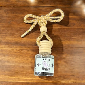 May include: A clear glass bottle with a wooden cap and a gold string hanging from it. The bottle has a label that says "Smoke Hash Eat Trash" and a cartoon image of a rodent smoking a joint.
