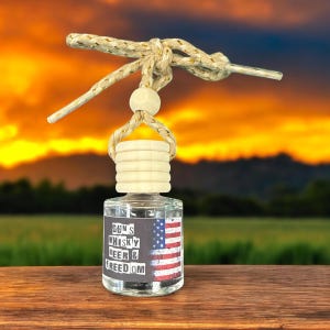 May include: A clear glass bottle car air freshener with a wooden cap and a string for hanging. The bottle has a black label with the text "Guns Whiskey Beer & Freedom" and a distressed American flag graphic.