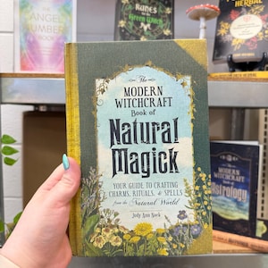 May include: A book titled "The Modern Witchcraft Book of Natural Magick" with a green and gold cover. The book is held in a hand and features floral illustrations and the author's name, Judy Ann Nock. Other books are visible in the background.