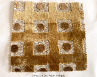 May include: A square fabric swatch of Cleopatra Cut Velvet in old gold featuring a grid pattern of raised velvet lines and circular velvet dots over a textured, light-colored woven base. The textile displays varying shades of gold and brown.
