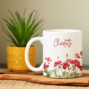 May include: White ceramic mug with a floral design featuring red geraniums. The mug has the name "Charlotte" written in pink script.