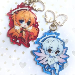 May include: Two acrylic keychains featuring anime-style characters. One keychain displays an orange-haired character in a red suit with a halo and spear. The other shows a white-haired character with wings and a halo, set against a blue background.