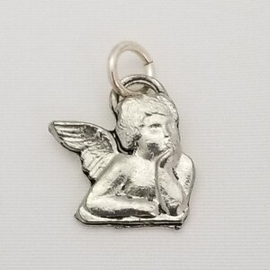 May include: Silver charm featuring a cherub angel with wings, sitting with its head resting on its hand.