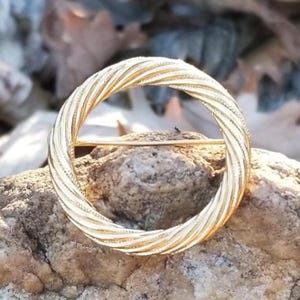 May include: A gold-colored circular brooch with a textured, twisted rope design. The brooch is set against a natural, textured stone background, highlighting its intricate details and metallic sheen. The pin is a classic accessory.