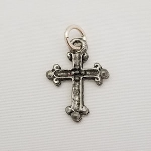 May include: A silver-toned cross charm with an ornate design. The cross has a loop at the top for attaching to a chain or bracelet.