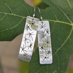 May include: A pair of rectangular silver earrings featuring a cut-out pattern of branching, star-like shapes. The metal surface has a hammered texture and is suspended from silver ear wires, creating a dangling, metallic design.