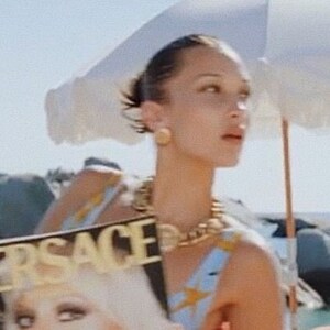 May include: A woman wearing a blue and yellow patterned swimsuit with a gold chain necklace holds a magazine with the word "VERSACE" printed on the cover. The magazine features a woman with blonde hair and makeup.