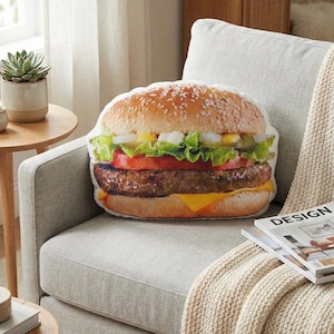 May include: A burger-shaped cushion featuring a sesame seed bun, lettuce, tomato, cheese, and a beef patty. The cushion rests on a light grey armchair with a cream blanket. A magazine is nearby.