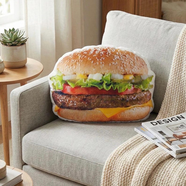May include: A burger-shaped cushion featuring a sesame seed bun, lettuce, tomato, cheese, and a beef patty. The cushion rests on a light grey armchair with a cream blanket. A magazine is nearby.