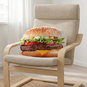 May include: A large burger-shaped cushion with sesame seed bun, salad, tomato, cheese, and a patty rests on a beige armchair. The chair has a light wood frame and neutral fabric. The burger cushion is the main feature.