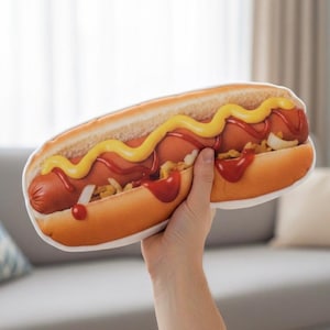 May include: A plush hot dog with a bun, sausage, mustard, ketchup, and onions. The hot dog is held up by a hand. The bun is light brown, the sausage is reddish-brown, and the condiments are yellow and red.