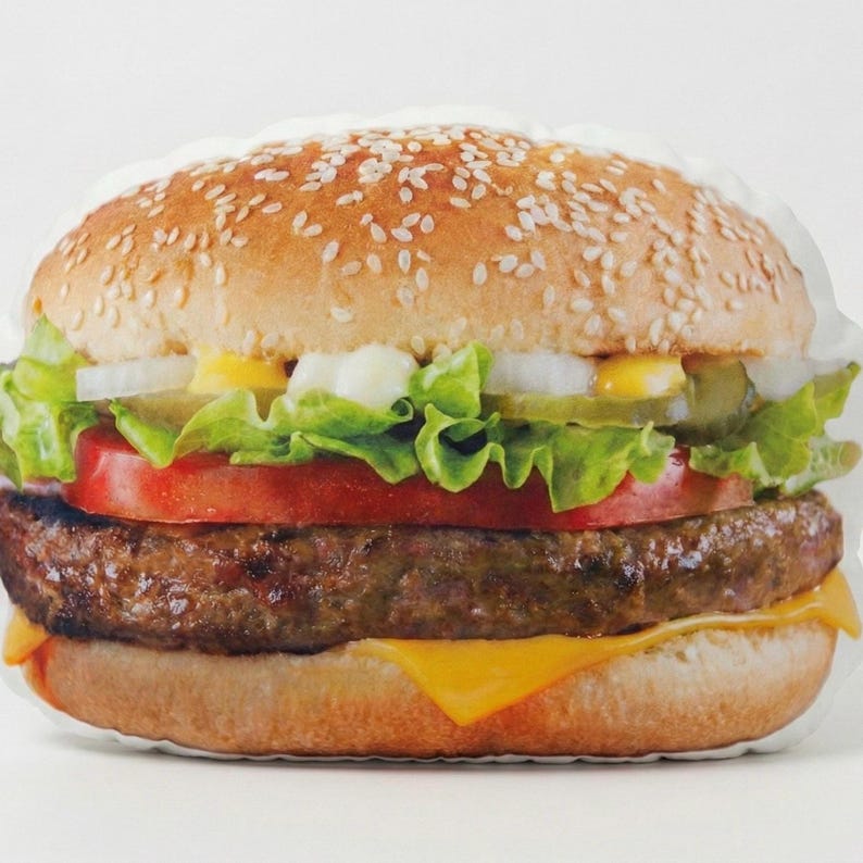 May include: A soft, cushion-like hamburger. The top bun is covered in sesame seeds, and the burger includes lettuce, tomato, onion, pickles, a beef patty, and melted cheese. It's designed to look like a classic cheeseburger.