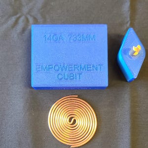 May include: A blue square and a blue diamond-shaped object with the text "14GA 733MM" and "EMPOWERMENT CUBIT" printed on the square. A copper spiral is also present. The objects are likely components of a larger item.