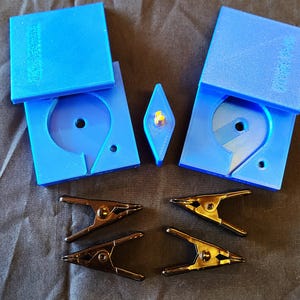 May include: An overhead shot shows a collection of crafting tools and accessories. Two open blue plastic cases with a circular cutout and a diamond-shaped object are displayed. Two pairs of metal spring clamps are also visible.