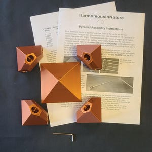 Pode incluir: Um kit de montagem de pirâmide de cor de cobre com instruções. O kit inclui quatro peças de canto, uma base quadrada e uma pequena chave hexagonal. As instruções são intituladas "Instruções de Montagem da Pirâmide" e incluem medidas.