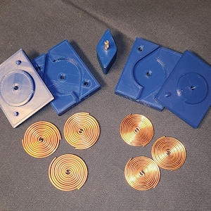 Puede incluir: Un conjunto de componentes, que incluye moldes de plástico azul y bobinas en espiral de cobre. Los moldes son cuadrados con hendiduras y agujeros circulares. También hay una pieza azul en forma de diamante con una inserción metálica dorada.