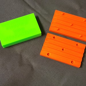 Op de afbeelding: Een stapel felgroene rechthoekige blokken en twee oranje rechthoekige blokken met een houtnerftextuur en gaten. De blokken liggen op een grijze ondergrond.