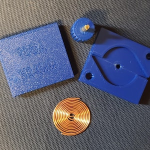 May include: A blue 3D printed device with a coil of copper wire, a small blue plastic piece with a hole in the center, and a blue plastic square with the text "18GA 0.84MM" printed on it.
