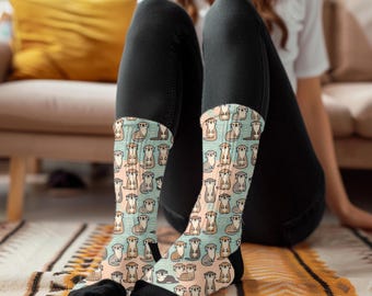 May include: A pair of socks with a pattern of cute otters on a light blue and pink background. The socks are worn by a person sitting on a rug.