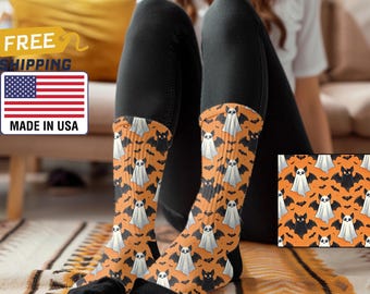 Puede incluir: Un par de calcetines naranja y negro con un patrón de fantasmas blancos y murciélagos negros. Los calcetines los lleva una persona sentada en una alfombra estampada.