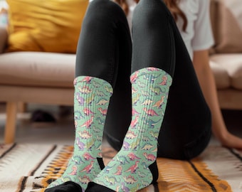 Puede incluir: Un par de calcetines con un estampado de dinosaurios coloridos sobre un fondo verde menta. Los calcetines los lleva puestos una persona sentada en un sofá.