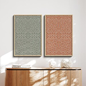 May include: Two framed abstract art prints. One print features a green and beige pattern, the other a red and white design. Both are rectangular with a repeating, organic pattern. The frames are a light wood color. The prints are displayed above a wooden cabinet.