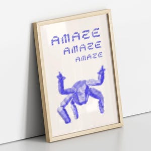 May include: A framed art print with the word "AMAZE" repeated three times in blue pixelated font. Below the text is a blue dotted graphic of a person with arms raised. The frame is light wood.