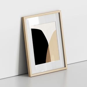 May include: A framed abstract art print featuring a minimalist design. The artwork showcases a black, beige, and tan color palette. The wooden frame has a natural finish, and the print is leaning against a white wall.