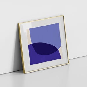 May include: Framed abstract art print featuring overlapping blue shapes in various shades. The artwork is displayed in a gold-colored frame with a white mat. The print leans against a white wall and a gray floor.