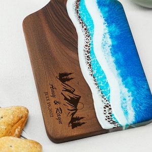 May include: A personalised wooden chopping board with a blue and white epoxy resin design resembling ocean waves. The board has a handle and is engraved with the text "Amy & Rayne JULY 19, 2022" and a mountain scene with pine trees.