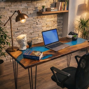 a wooden desk with a laptop, a lamp, a book, and a statue on it. The desk is situated in a room with a stone wall and a shelf with books. There is also a black chair and a potted plant in the room.
