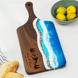 May include: A wooden serving board with a handle, featuring a dark wood grain finish. The right side has a blue and white resin design resembling ocean waves. The board is engraved with text and a mountain scene.