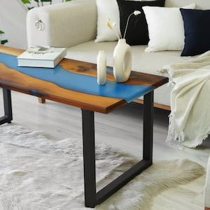 Puede incluir: Una mesa de centro de madera con un diseño de río de epoxi azul. La mesa tiene patas de metal negro y está sobre una alfombra blanca y esponjosa.
