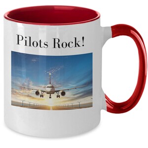 May include: White ceramic coffee mug with a red rim and handle. The mug features a graphic of an airplane landing on a runway with a blue sky and clouds in the background. The text "Pilots Rock!" is printed on the mug.
