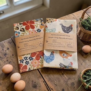 Puede incluir: Dos envoltorios de cera de abeja hechos a mano con estampados florales y de gallinas, de 43x53 cm, se exhiben sobre una mesa de madera. Los envoltorios están etiquetados como "Simple Living, Naturally Wrapped". También hay varios huevos y una cesta de hierbas.