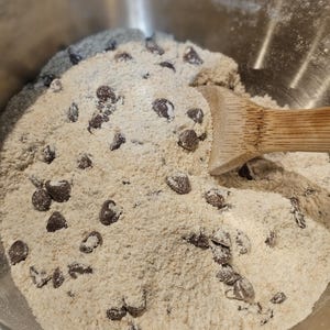 Puede incluir: Primer plano de un cuenco de acero inoxidable lleno de ingredientes para masa de galletas. La masa es de color marr&oacute;n claro, salpicada de chispas de chocolate negro. Una cuchara de madera descansa en la masa.